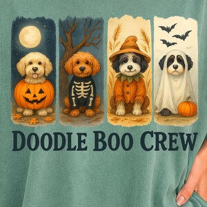 May include: A sage green t-shirt featuring a graphic of four dogs in Halloween costumes with the text "DOODLE BOO CREW" underneath. The dogs are depicted as a pumpkin, a skeleton, a witch, and a ghost, set against a seasonal autumn background.