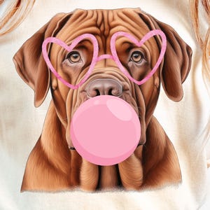 May include: Cream-colored t-shirt featuring a brown dog with pink heart-shaped glasses and a pink bubblegum bubble. The dog's face is the focal point, with detailed fur and expressive eyes. The shirt is a casual style.