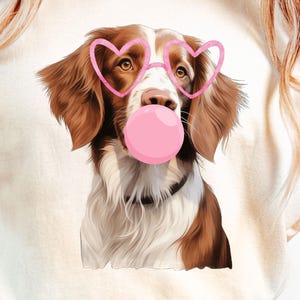 Pode incluir: Camiseta creme com um cachorro com pelo marrom e branco, usando óculos rosa em forma de coração e uma bolha de chiclete rosa. O retrato do cachorro está centralizado na camiseta.