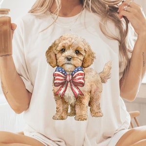 May include: A white t-shirt featuring a graphic of a brown and white dog wearing a red, white and blue bow. The dog is sitting on a white background.