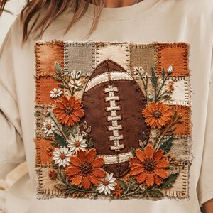 Könnte beinhalten: Ein beiges T-Shirt mit kurzen Ärmeln und einem zentralen Motiv eines braunen Footballs, der von orangefarbenen und weißen Blumen umgeben ist. Das Design ist auf einem Hintergrund aus Patchwork-Quadraten in Braun-, Orange- und Grüntönen platziert.