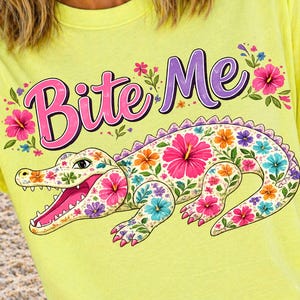May include: Yellow t-shirt with the text "Bite Me" in pink and purple, decorated with flowers. A floral alligator design in pink, purple, blue, and orange is featured below the text. The shirt is worn on a beach.