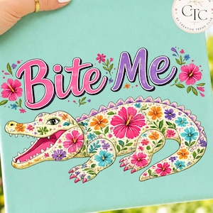 May include: A canvas print with a floral alligator design and the words "Bite Me". The alligator is decorated with colourful flowers in shades of pink, orange, and blue. The background is a light teal colour. The design is by Creative Trend Club.
