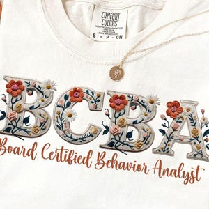 Pode incluir: Uma t-shirt de cor creme com as letras BCBA num estilo bordado floral, com o texto Board Certified Behavior Analyst impresso por baixo numa fonte cursiva castanha. A t-shirt tem uma etiqueta da marca Comfort Colors no decote.