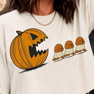 May include: An off-white short-sleeved t-shirt featuring a graphic of a large orange jack-o'-lantern with a jagged mouth chasing three brown dogs wearing white ghost costumes. The design is printed on the front of the garment.