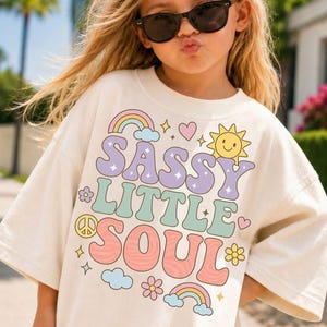 May include: An off-white short-sleeve t-shirt featuring a colorful graphic print with the text SASSY LITTLE SOUL in wavy, retro-style lettering. The design includes illustrations of rainbows, a smiling sun, a peace sign, hearts, and flowers.
