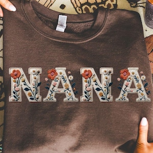 May include: A brown sweatshirt with the word "NANA" embroidered across the front. The letters are white with floral designs in shades of orange, yellow, and green. The sweatshirt is held by two hands.