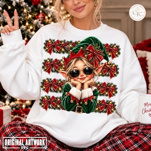 Peut inclure: Un sweat-shirt blanc à manches longues avec une impression graphique représentant un elfe portant des lunettes de soleil et un chapeau vert. Le motif comprend des rangées horizontales de guirlandes décorées de nœuds rouges et de clochettes dorées.