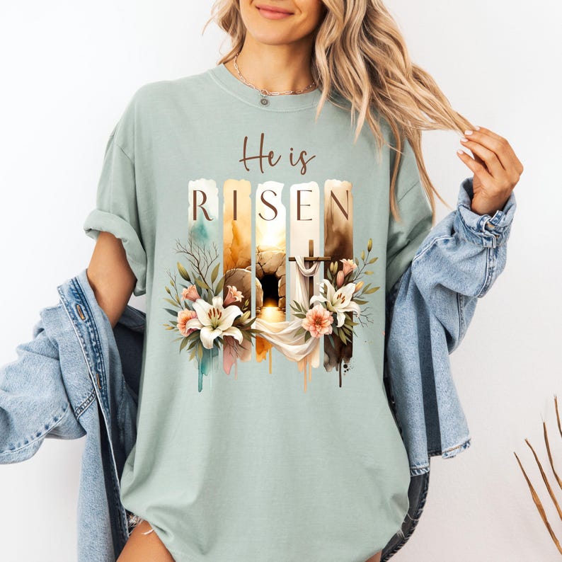 Puede incluir: Una camiseta azul claro con un dise&ntilde;o de acuarela de una tumba con una cruz y flores. El texto "He is Risen" est&aacute; impreso en la camiseta.