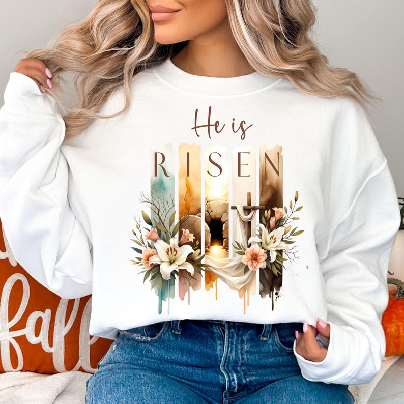 Puede incluir: Sudadera blanca con un dise&ntilde;o de acuarela de una tumba, flores y una cruz. El texto "He is Risen" est&aacute; impreso en la sudadera.