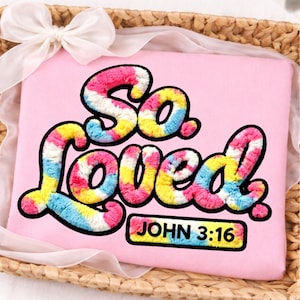 May include: A pink square pillow with the words "So Loved" in colorful, textured lettering. The text "JOHN 3:16" is in a small rectangle at the bottom. The pillow is in a woven basket with a white ribbon.