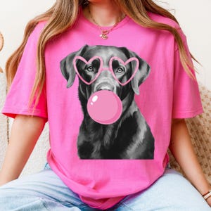 May include: A pink t-shirt featuring a black and white illustration of a Labrador Retriever wearing pink heart-shaped glasses and blowing a pink bubble. The dog's image is centered on the shirt.