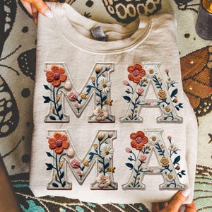 May include: Cream-colored sweatshirt with the word "MA" embroidered twice in floral letters. The letters are adorned with orange, pink, and white flowers, and green leaves. The sweatshirt is folded and held by hands.