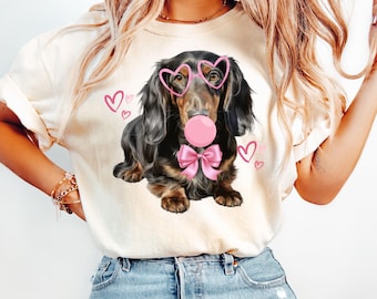 Puede incluir: Una camiseta blanca con un dachshund negro y marrón que lleva gafas de sol en forma de corazón rosa y un lazo rosa. El dachshund está soplando una burbuja rosa y hay corazones rosas en el fondo.