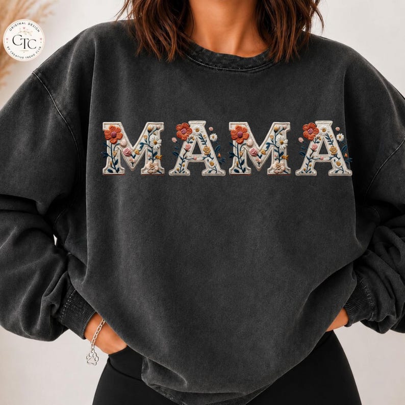May include: A charcoal grey crewneck sweatshirt featuring the word MAMA printed across the chest. Each letter is filled with a detailed floral pattern consisting of orange, yellow, white, and pink flowers with green leaves and stems on a cream background.