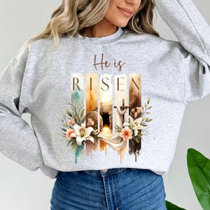 Puede incluir: Una sudadera gris con las palabras "He is Risen" impresas en la parte delantera. El texto est&aacute; rodeado por un dise&ntilde;o de acuarela de una tumba, una cruz y flores.