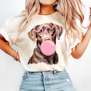 May include: A white t-shirt with a brown dog wearing pink heart-shaped sunglasses and blowing a pink bubble. The dog is looking directly at the camera.