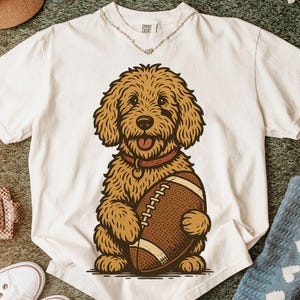 May include: A white short-sleeved t-shirt featuring a graphic illustration of a golden-brown curly-haired dog wearing a red collar and holding a brown football. The shirt has a crew neckline and a Comfort Colors brand label at the inner neck.