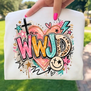 Könnte beinhalten: Weißes Sweatshirt mit einem farbenfrohen Grafikdesign. Das Design enthält die Buchstaben "WWJD" in Pink, Orange und Türkis, ein Kreuz, Sterne, Herzen und ein Smiley. Der Hintergrund hat einen Aquarell-Effekt.