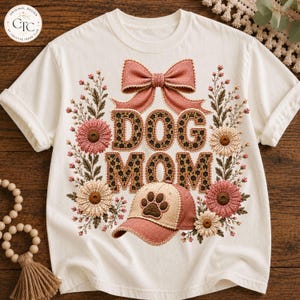 May include: A cream-coloured t-shirt featuring a graphic design with the text "DOG MOM" in a leopard print pattern. The design includes a large pink bow above the text, a baseball cap with a paw print below, and floral accents in pink and beige.