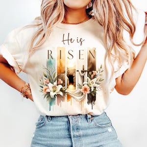 Puede incluir: Una camiseta blanca con un dise&ntilde;o de acuarela que presenta una tumba, una cruz y flores. El texto "He is RISEN" est&aacute; impreso en la camiseta.