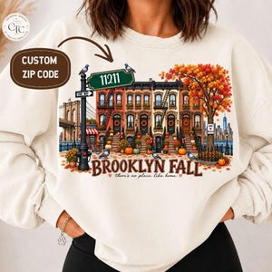 May include: A cream-coloured crewneck sweatshirt featuring a graphic print of Brooklyn brownstone buildings in autumn. The design includes a street sign reading 11211, a tree with orange leaves, pumpkins, pigeons, and the text BROOKLYN FALL there's no place like home.