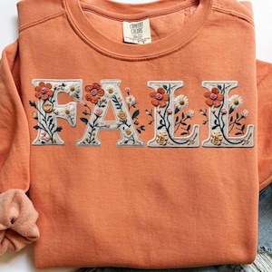 May include: An orange sweatshirt with the word "FALL" embroidered across the front. The letters are white with floral embroidery in shades of orange, yellow, and green. The sweatshirt has a crew neck and long sleeves.