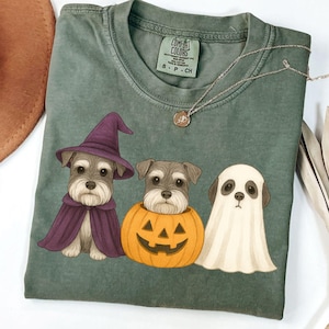 Puede incluir: Una camiseta verde Comfort Colors con un diseño de Halloween. Presenta tres perros Schnauzer: uno con disfraz de bruja, otro en una calabaza y otro como fantasma. También se ve un collar plateado con un colgante.