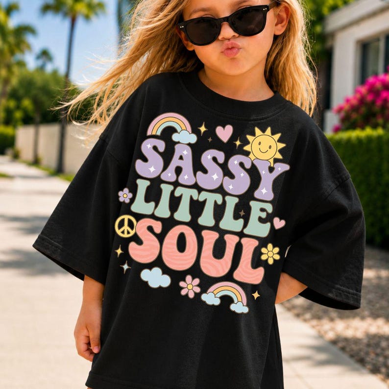 May include: A black t-shirt featuring the text SASSY LITTLE SOUL in large, wavy, multi-colored letters. The design includes graphic elements such as rainbows, a smiling sun, hearts, flowers, a peace sign, clouds, and small stars in pastel tones.