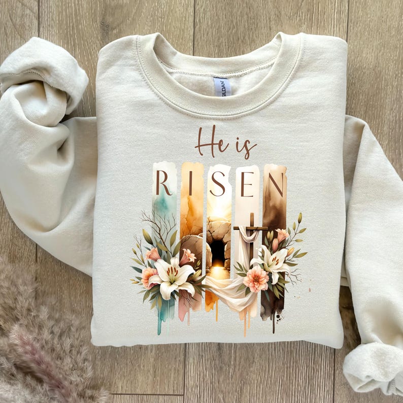 Puede incluir: Una sudadera beige con un dise&ntilde;o de acuarela de una tumba, una cruz y flores. El texto "He is RISEN" est&aacute; impreso en la sudadera.