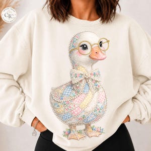 Puede incluir: Una sudadera de cuello redondo color crema con un gráfico central de un pato con gafas de montura dorada. El pato está decorado con un patrón de patchwork de cuadrados florales y de cuadros vichy en azul, rosa y amarillo pastel, con un gran lazo.