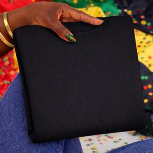 May include: A folded black crew neck sweatshirt. The sweatshirt is held by a hand with gold bracelets and decorated nails. The background features a colorful pattern of red, green, yellow, and blue.