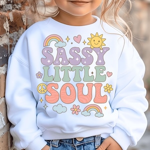 May include: A white long-sleeve sweatshirt featuring a colorful graphic print with the text SASSY LITTLE SOUL. The design includes pastel-colored rainbows, a smiling sun, flowers, a peace sign, hearts, and clouds in shades of pink, yellow, and blue.