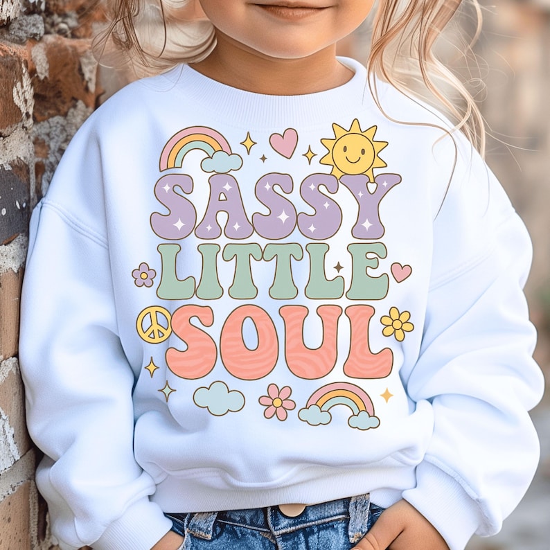 May include: A white long-sleeve sweatshirt featuring a colorful graphic print with the text SASSY LITTLE SOUL. The design includes pastel-colored rainbows, a smiling sun, flowers, a peace sign, hearts, and clouds in shades of pink, yellow, and blue.