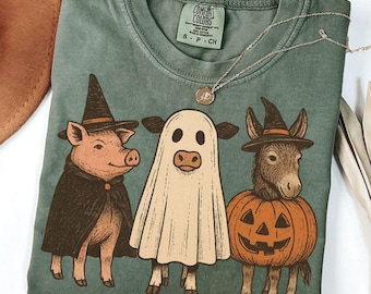 May include: Green Comfort Colors t-shirt featuring a Halloween-themed graphic. The graphic includes a pig in a witch hat and cape, a cow in a ghost costume, and a donkey in a pumpkin costume. A silver necklace with a pendant is also visible.