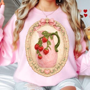 Op de afbeelding: Roze sweatshirt met een vintage-stijl illustratie van een roze kan met rode aardbeien en groene bladeren. De kan staat in een ovale lijst met een bloemen- en kantontwerp, bekroond met een roze strik. Onderaan een hartvormig detail.