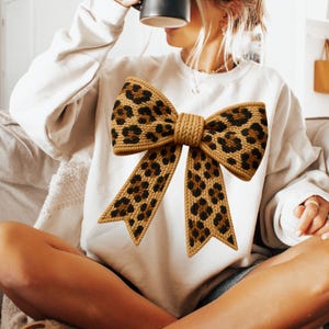 Puede incluir: Sudadera color crema con un gran diseño de lazo. El lazo tiene un estampado de leopardo con manchas marrones y negras, y un nudo central de color beige. Es una prenda casual para el uso diario.
