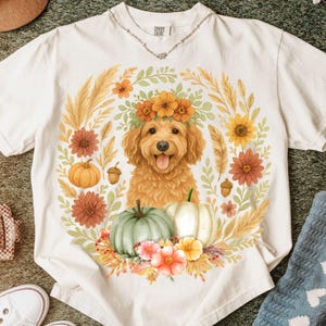 May include: A cream-colored short-sleeve t-shirt featuring a central graphic of a golden-brown dog with a floral crown, surrounded by a wreath of autumn flowers, wheat stalks, acorns, and two pumpkins in shades of orange and sage green.