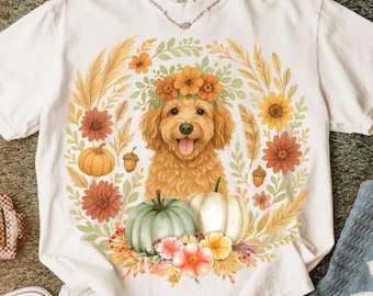 May include: A cream-colored short-sleeve t-shirt featuring a central graphic of a golden-brown dog with a floral crown, surrounded by a wreath of autumn flowers, wheat stalks, acorns, and two pumpkins in shades of orange and sage green.