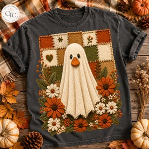 Könnte beinhalten: Ein anthrazitfarbenes T-Shirt mit einer zentralen Grafik einer geisterhaften Ente, die in ein weißes Laken gehüllt ist, vor einem Patchwork-Hintergrund aus herbstfarbenen Quadraten, umgeben von orangefarbenen und weißen Häkelblumen.