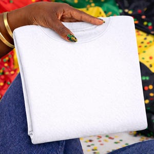 May include: A folded white crew neck sweatshirt. The sweatshirt is held by a hand with gold bracelets and decorated nails. The background includes colorful fabric and confetti.