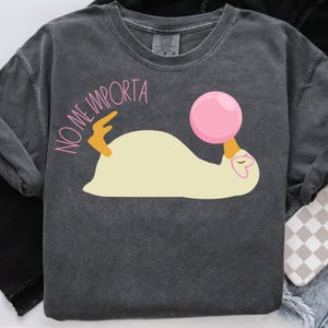 Puede incluir: Una camiseta gris oscura con un pato blanco y una pelota rosa. El texto "NO ME IMPORTA" está impreso en rosa en la camiseta.