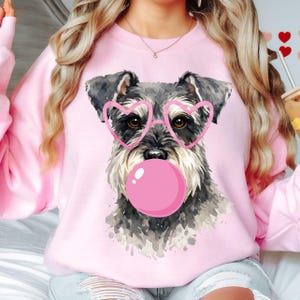 May include: A light pink crewneck sweatshirt featuring a watercolor-style illustration of a Schnauzer dog wearing pink heart-shaped glasses and blowing a large, round, bright pink bubblegum bubble centered on the front of the garment.
