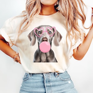 Könnte beinhalten: Ein weißes T-Shirt mit einem Aufdruck eines grauen Hundes, der eine rosafarbene Herzbrille trägt und eine rosafarbene Blase bläst.