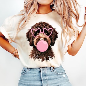 May include: A brown and white dog wearing pink heart-shaped sunglasses and blowing a pink bubble. The dog is on a white t-shirt.