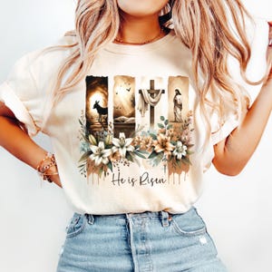 May include: Cream-colored t-shirt featuring a religious graphic. The design includes four vertical panels depicting scenes with a donkey, empty tomb, cross, and Jesus. Floral accents and the text "He is Risen" complete the design.