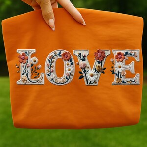 May include: Orange tote bag with the word "LOVE" embroidered in white, each letter adorned with colorful floral designs. The flowers include daisies, roses, and other blooms in shades of orange, pink, and green.