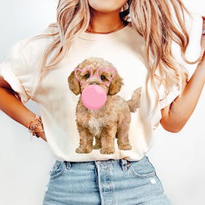 Puede incluir: Una camiseta blanca con un gráfico de un perro marrón y blanco que lleva gafas de corazón rosa y sopla una burbuja rosa.