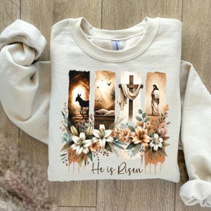 May include: Cream-colored sweatshirt featuring a religious graphic with a cross, Jesus, and the text "He is Risen." The design includes floral elements and a donkey. The sweatshirt is made of a soft material.