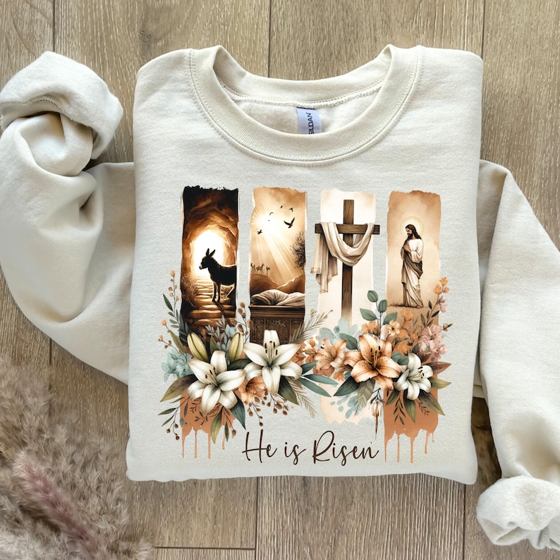 May include: Cream-colored sweatshirt featuring a religious graphic with a cross, Jesus, and the text "He is Risen." The design includes floral elements and a donkey. The sweatshirt is made of a soft material.